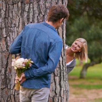 Romance in the park. A young man hiding flowers behind his back while his girlfriend jumps out behind a tree