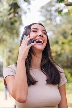 young caucasian woman laughing during a call with her mobile phone in a park, concept of youth and technology of communication