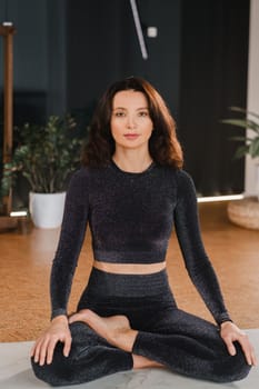 A woman in a black tracksuit sits on a yoga mat in a lotus position in the gym.