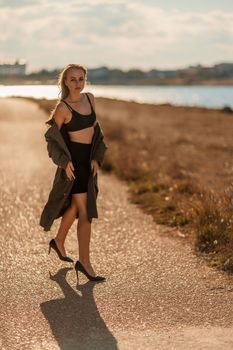 Portrait blonde sea cape. A calm young blonde in an unbuttoned khaki raincoat walks along the seashore, under a raincoat a black skirt and top.