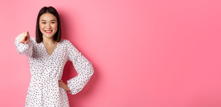 Confident asian woman in dress pointing at you, smiling and staring at camera, standing against pink background. Copy space