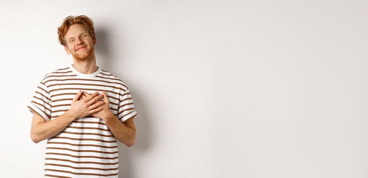 Happy young man with ginger hair appreciate gift, holding hands on heart and smiling, thanking you, standing grateful against white background.