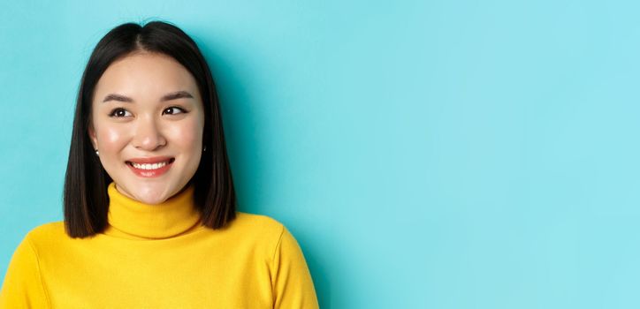 Beauty and makeup concept. Dreamy asian girl with perfect healthy skin, looking left and smiling happy, standing over blue background.