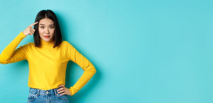 Are you stupid. Portrait of skeptical asian woman scolding someone crazy or dumb, pointing finger at head and staring at camera with dismay, blue background.