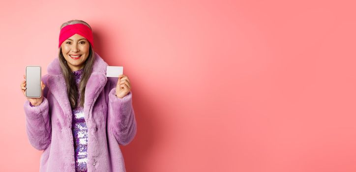 Online shopping and fashion concept. Fashionable asian senior woman showing black screen of smartphone and plastic credit card, smiling happy, pink background.