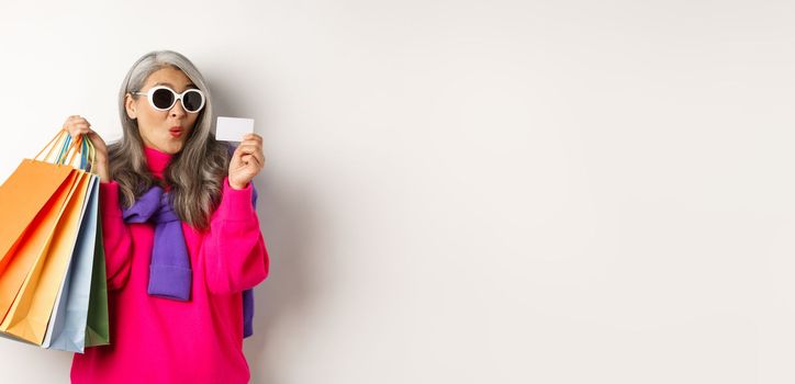 Fashionable asian old lady going shopping, holding paper bags and plastic credit card, wearing stylish sunglasses, standing over white background.