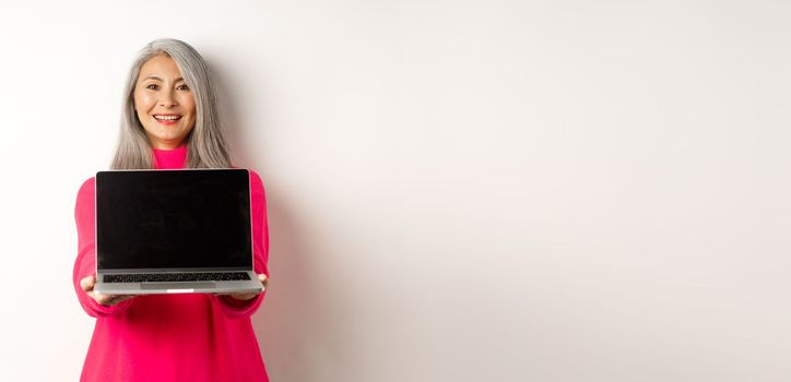 E-commerce concept. Smiling asian senior woman showing blank laptop screen and looking happy, demonstrating promo, white background.