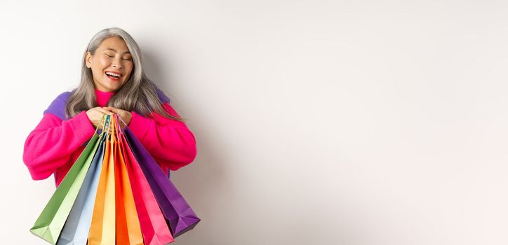 Fashionable senior asian woman shopaholic, hugging shopping bags and smiling joyful, buying with discounts, standing over white background.