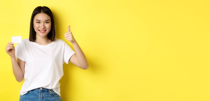 Young asian woman in casual white t-shirt showing plastic credit card and thumb up gesture, approve and recommend, smiling at camera, yellow background.