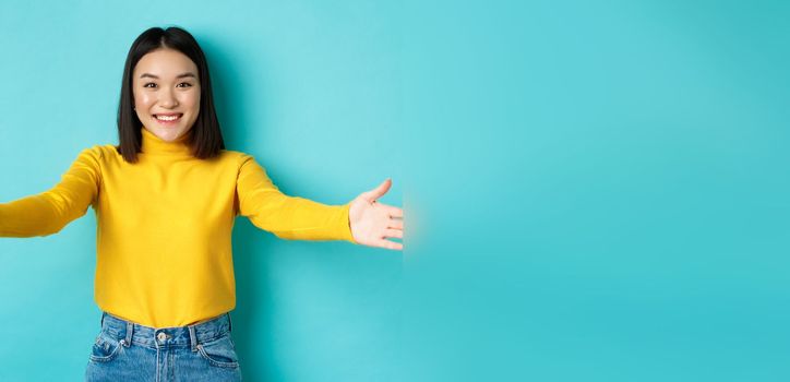 Portrait of beautiful korean woman spread out hands for hug, reaching for cuddles and smiling at camera, greeting you, standing over blue background.