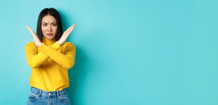 Serious and assertive korean woman showing cross stop gesture, frowning and telling no, prohibit action, disapprove something bad, standing over blue background.