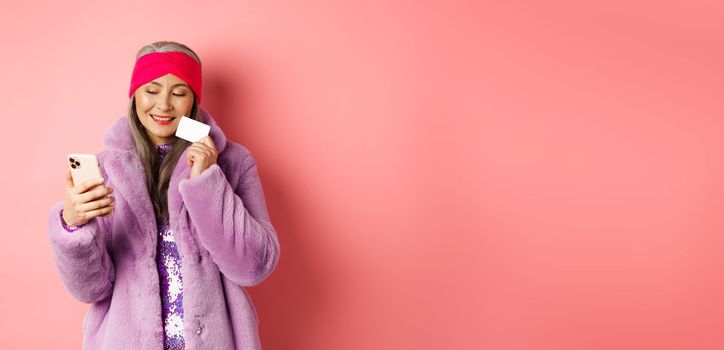 Online shopping and fashion concept. Stylish asian grandmother making purchase in internet with smartphone and plastic credit card, pink background.