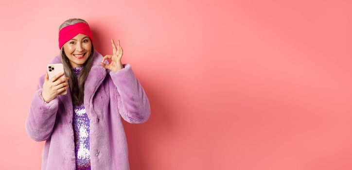 Online shopping and fashion concept. Stylish asian senior woman showing okay sign and holding mobile phone, recommending internet store, pink background.