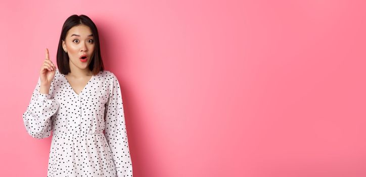 Excited asian girl having an idea, saying suggestion, raising finger and talking, standing over pink background.