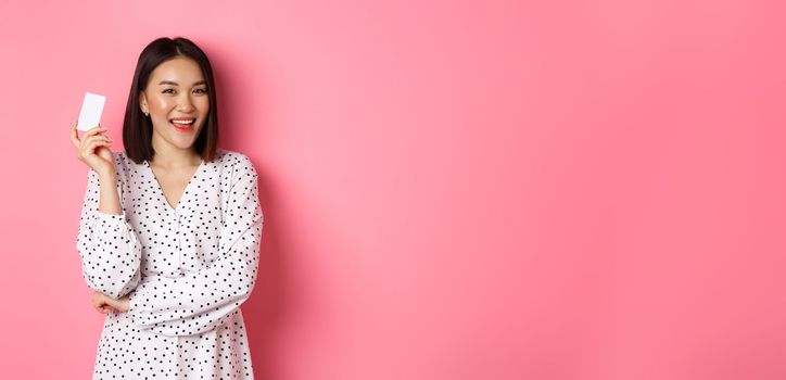 Shopping concept. Confident and happy asian woman holding credit card and smiling satisfied, standing against pink background.