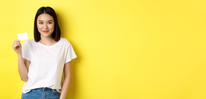 Young asian woman in casual white t-shirt showing plastic credit card and smiling at camera, yellow background.