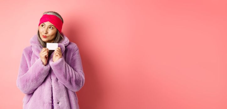 Shopping and fashion concept. Fashionable asian senior woman thinking about buying gifts, holding credit card and looking thoughtful at upper left corner, pink background.