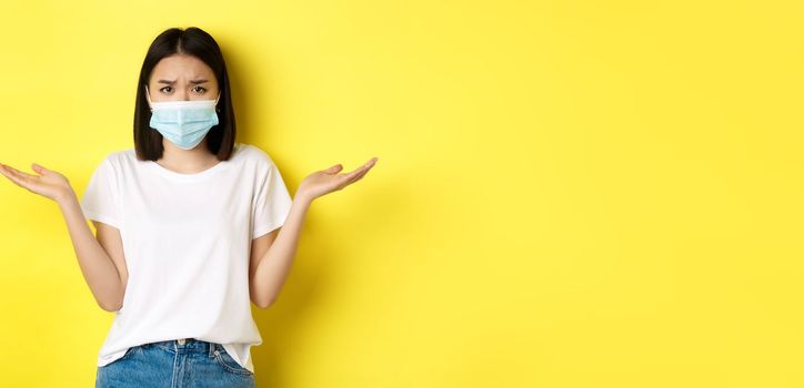 Covid-19, quarantine and social distancing concept. Confused asian woman in medical mask shrugging shoulders, spread hands sideways clueless, know nothing, yellow background.