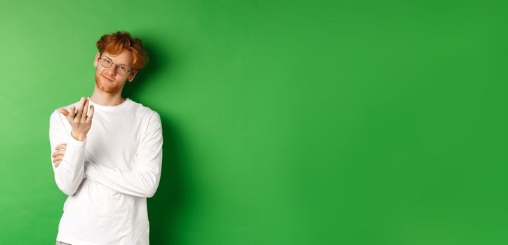 Young man with red messy hair and glasses, smiling pleased and pointing hand at camera, your turn gesture, standing over green background.