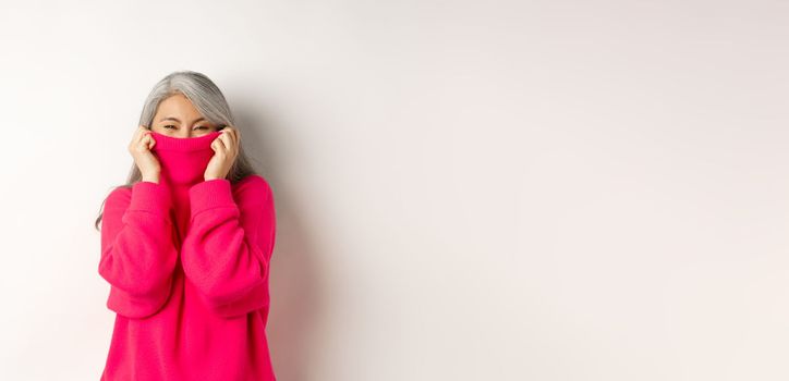 Cheerful asian senior woman laughing, wearing warm sweater, hiding face inside collar, standing happy over white background.