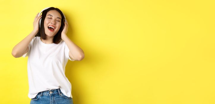 Attractive asian woman enjoying listening music in wireless headphones, smiling pleased and singing along, yellow background.