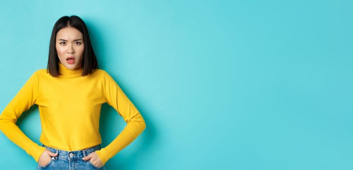 Portrait of disappointed korean woman in yellow sweater, frowning and looking upset, standing over blue background.