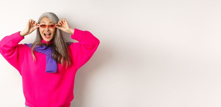 Fashionable asian senior woman trying new sunglasses, looking amazed and cheerful at camera, standing in trendy sweater against white background.