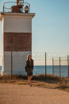 Portrait blonde sea cape. A calm young blonde in a khaki raincoat stands on the seashore against the backdrop of a lighthouse