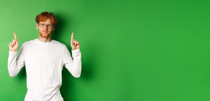 Displeased young man with red hair and glasses, pointing fingers up and frowning doubtful, feeling disappointed, standing over green background.