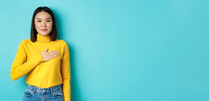 Image of proud smiling asian woman holding hand on heart, showing respect to national anthem, standing over blue background.