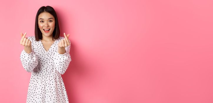 Lovely asian woman in dress showing korean heart signs and smiling, standing on romantic pink background.