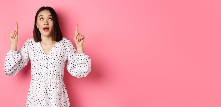 Image of beautiful asian girl in dress looking, pointing fingers up, gazing dreamy at promo offer, standing over pink background.