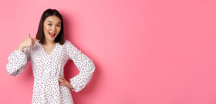 Beautiful asian girl in dress looking happy, pointing finger at herself, standing on romantic pink background.