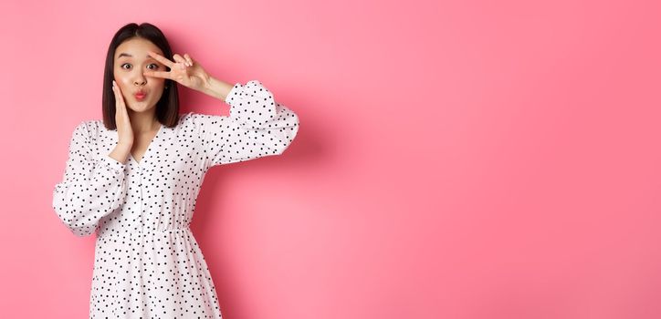 Coquettish asian woman in dress showing peace sign on eye, pucker lips for kiss, standing silly against pink background.