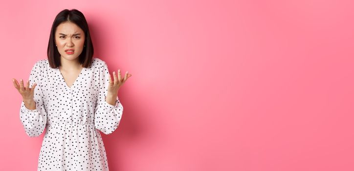 Angry and confused asian woman pointing hands at camera and grimacing furious, standing annoyed against pink background.