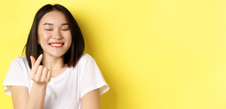 Valentines day and women concept. Close up of pretty asian girl in white t-shirt, smiling and showing finger heart, I love you gesture, standing over yellow background.