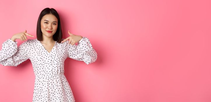 Beautiful asian woman looking confident, pointing at herself and smiling, showing logo on chest, standing over pink background.