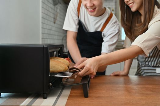 Cropped young couple taking off french baguette bread from oven, preparing dinner in modern kitchen together.