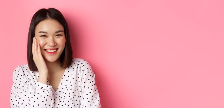 Close-up of adorable asian woman blushing, touching cheek and smiling cute at camera, standing over pink background.