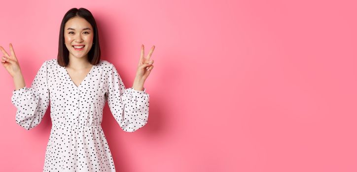 Cute asian brunette girl in dress smiling, showing kawaii peace signs and looking happy, standing over pink background.