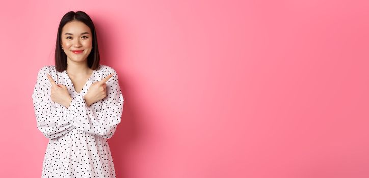 Beautiful asian girl in romantic dress pointing fingers sideways, showing two variants on shopping, smiling at camera, standing against pink background.