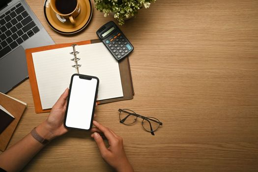 Overhead view of woman hand holding smart phone with white blank screen over wooden office desk.