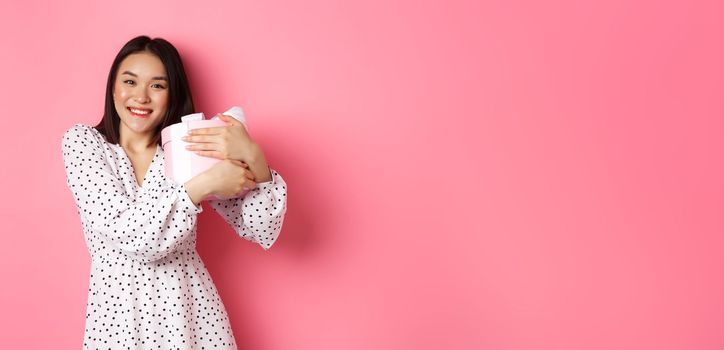 Lovely asian woman hugging her gift and smiling thankful, receive valentines day present, standing over pink background.