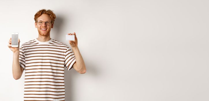 Shopping and finance concept. Young man showing blank mobile screen and plastic credit card, smiling at camera, white background.