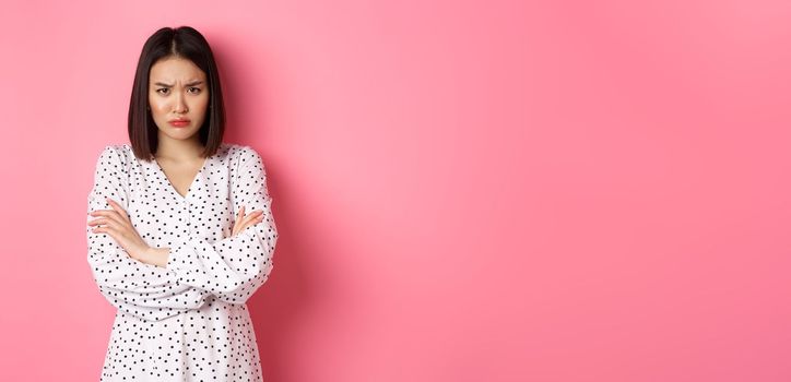 Timid and offended asian girl cross arms on chest, staring defensive and insulted at camera, standing in dress over pink background.