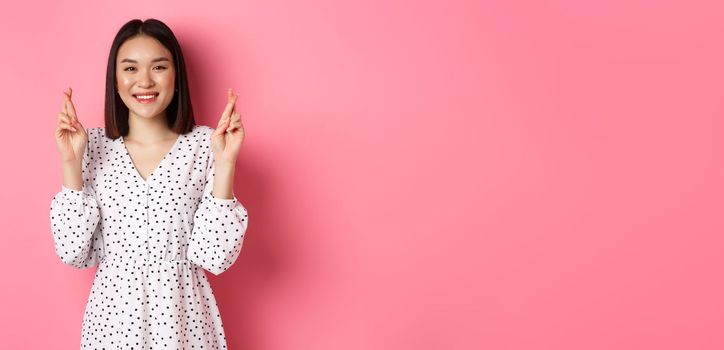 Cute asian woman making a wish, smiling happy and cross fingers for good luck, waiting over pink background.