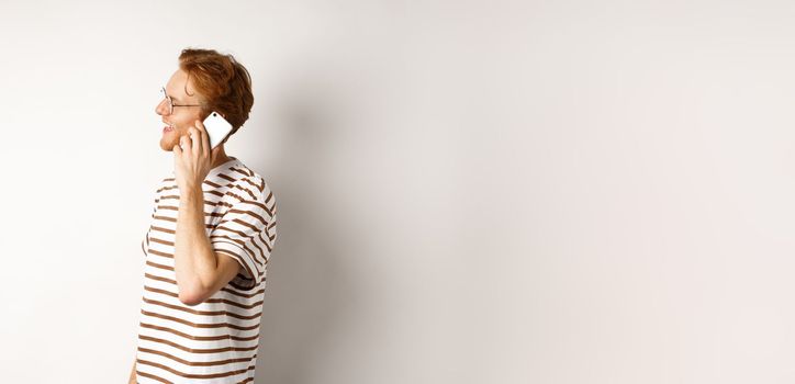 Profile of happy young handsome man talking on mobile phone, looking left and smiling, standing over white background.