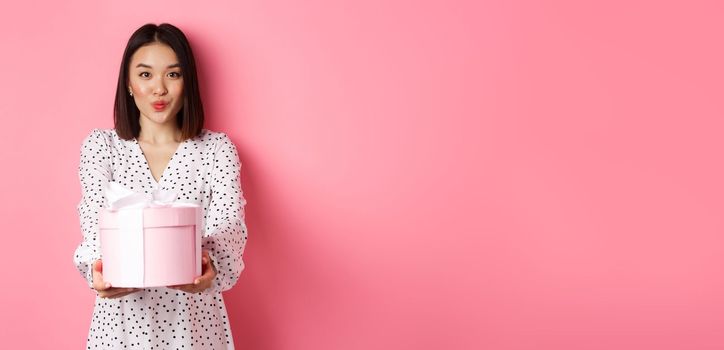 Cute asian girlfriend congratulate with valentines day, giving cute romantic gift in box, pucker lips for kiss, standing over pink background.