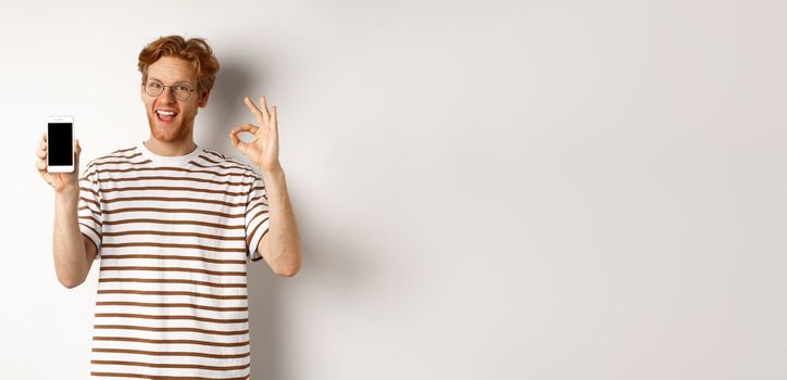 Technology and e-commerce concept. Young man with red hair showing okay sign and blank smartphone screen, praising awesome app, standing over white background.