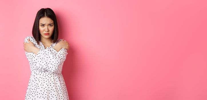 Timid and offended asian girl cross arms on chest, staring defensive and insulted at camera, standing in dress over pink background.